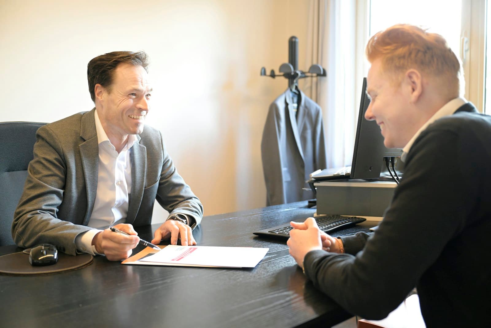 A professional in a blazer smiles at a client across an office desk, pen in hand over a document.