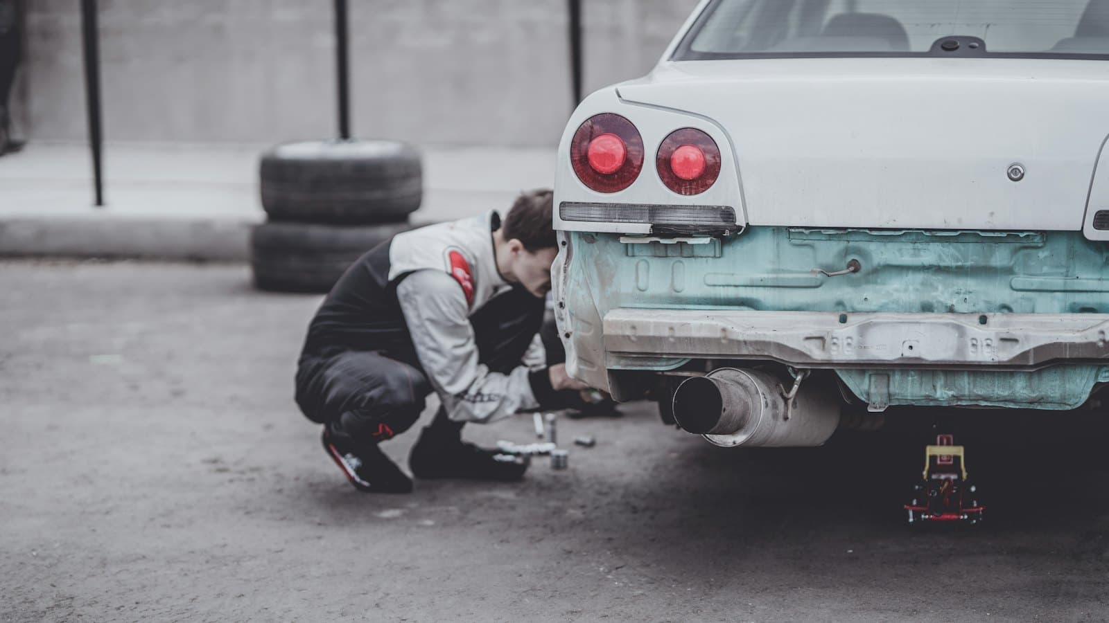 A technician crouches behind a white car with its rear bumper removed, working on the bare body panel.