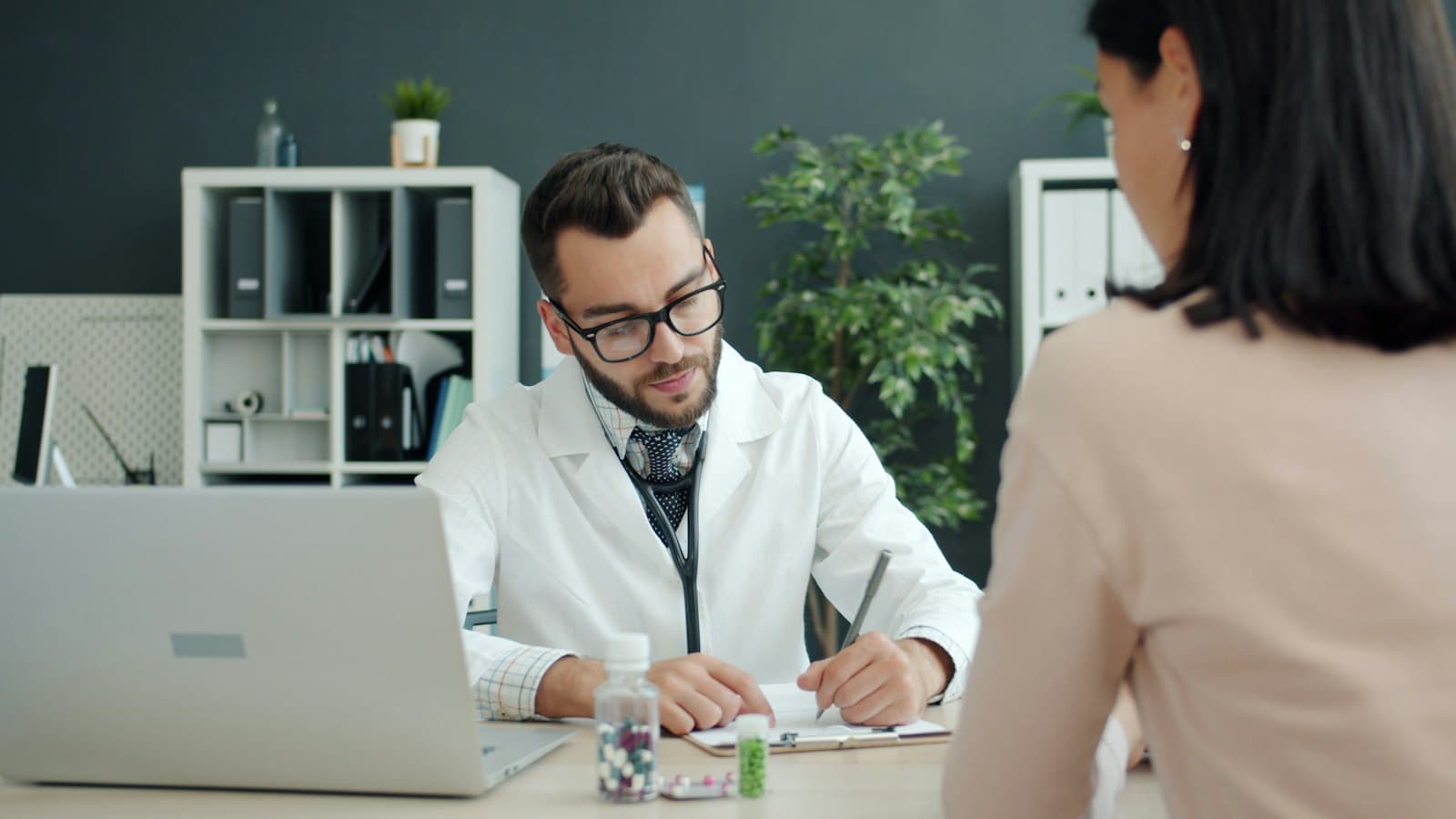 A doctor in a white coat takes notes at a desk across from a patient, who is seen from behind.