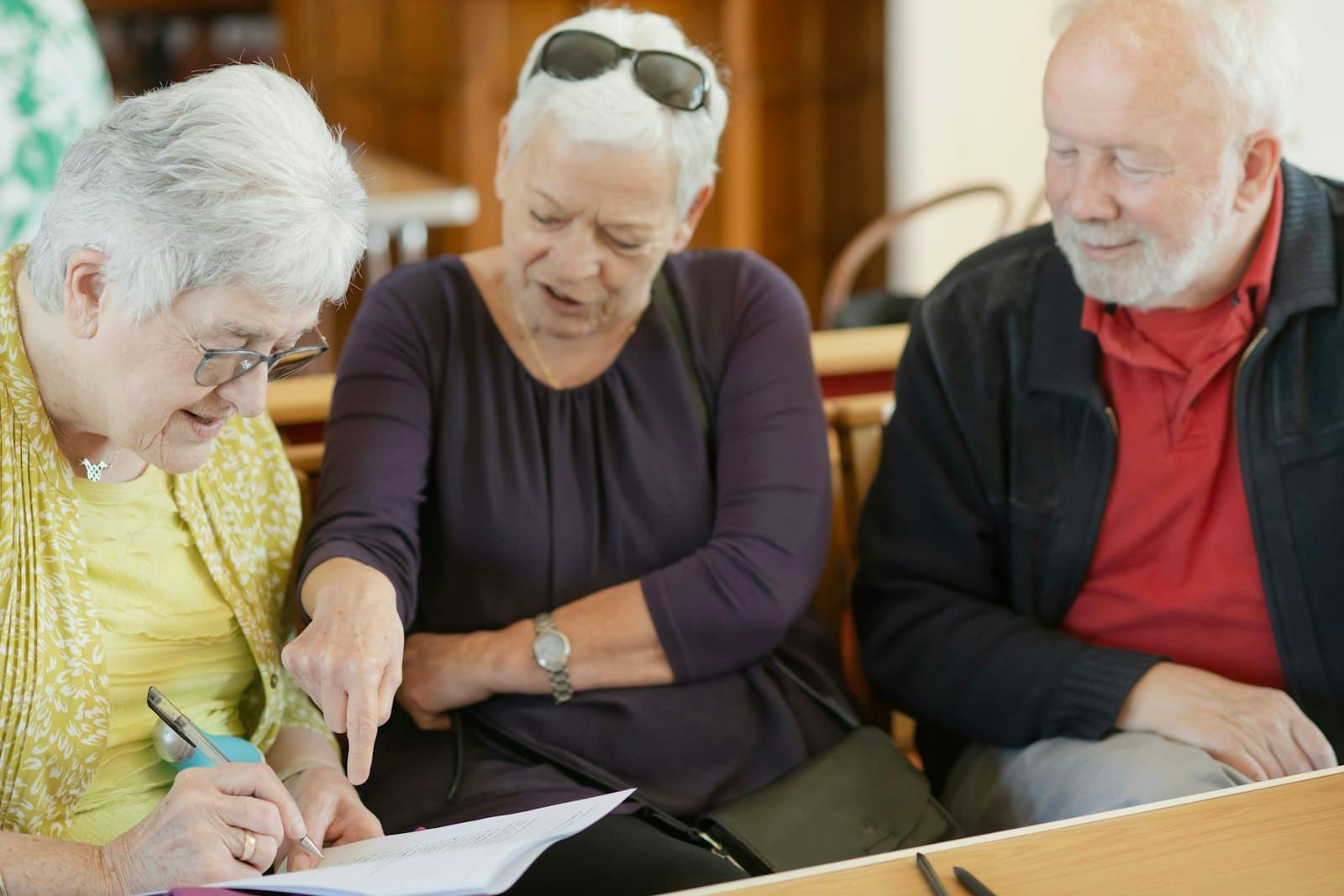 A woman points to a form while two older adults look on and one fills it in at a table.