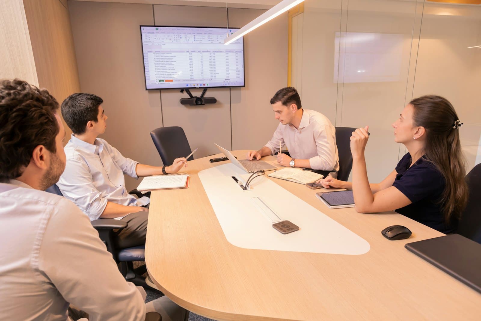 A small team of four meets around a conference table, looking up at a spreadsheet on a wall screen.