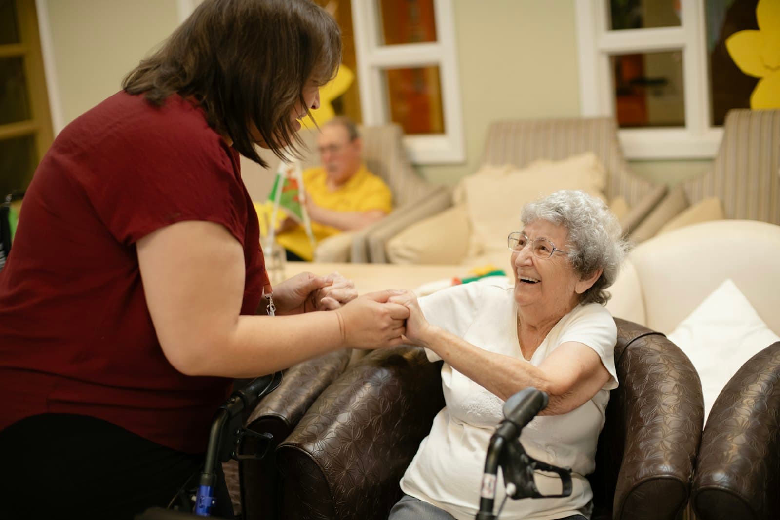 A caregiver holds the hands of a smiling older woman as she rises from her chair with a walker.