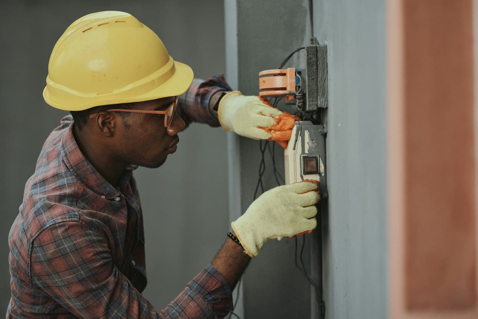 An electrician in a yellow hard hat and work gloves wires an electrical box on a wall.