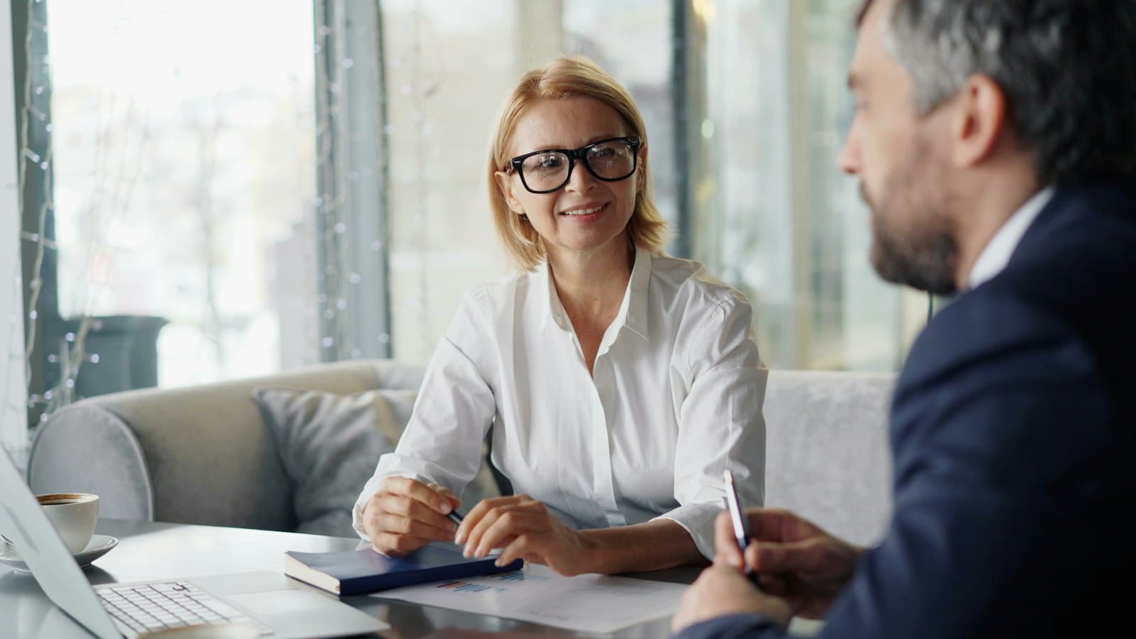 A smiling woman in glasses meets with a client across a desk with a laptop and notebook.