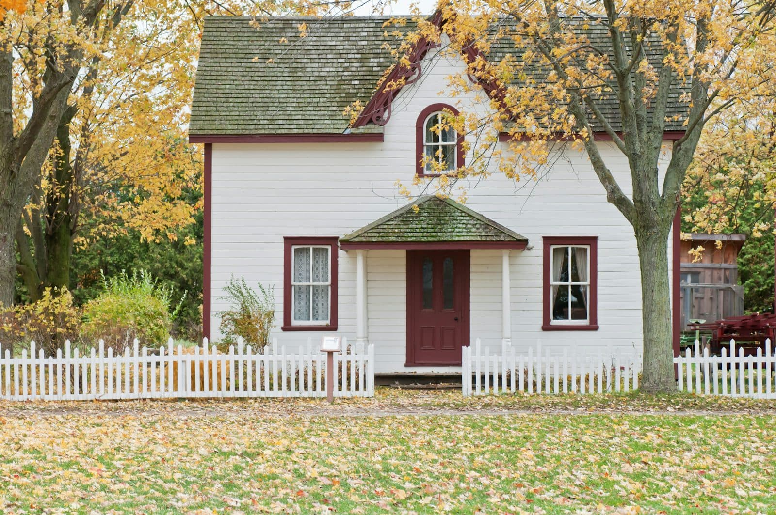 A white two-story house with a red front door and a white picket fence, framed by autumn trees and a leaf-covered lawn.