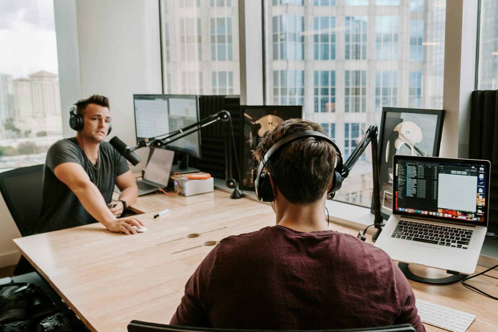 Two people in headphones talk into boom-arm microphones across a wooden table by a sunny window.