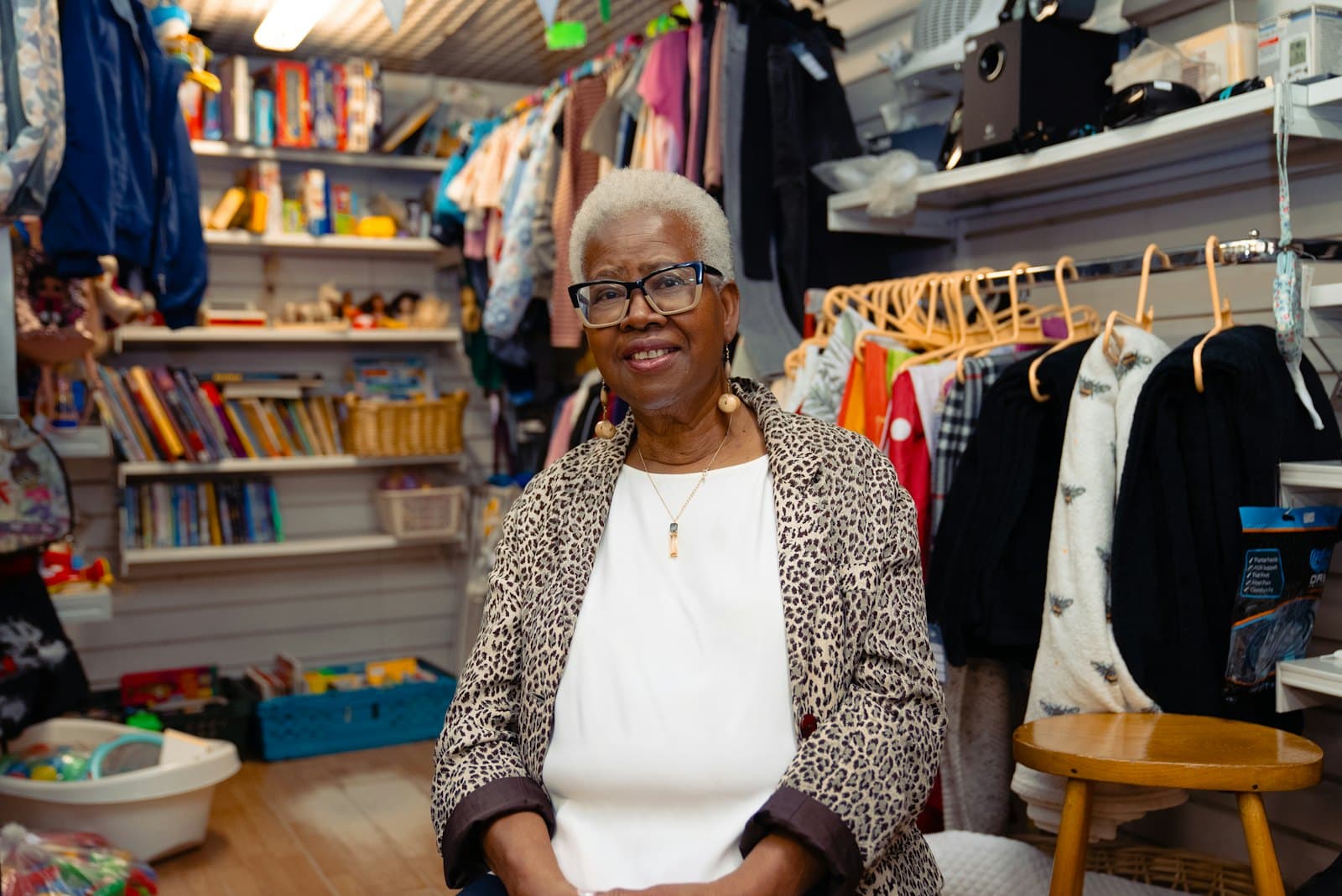 An older woman in glasses smiles inside her small shop, surrounded by racks of clothes, books and toys.