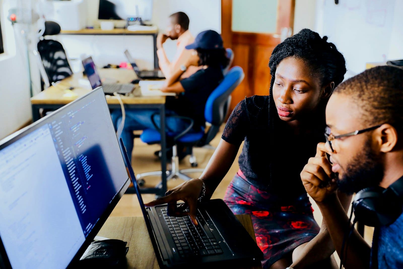 A woman points at a laptop screen to help a colleague in a small, busy office.